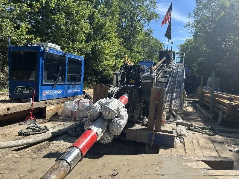 Drilling rig on a construction site with a red drill pipe and nearby equipment, surrounded by trees and a blue sky flag in the background.