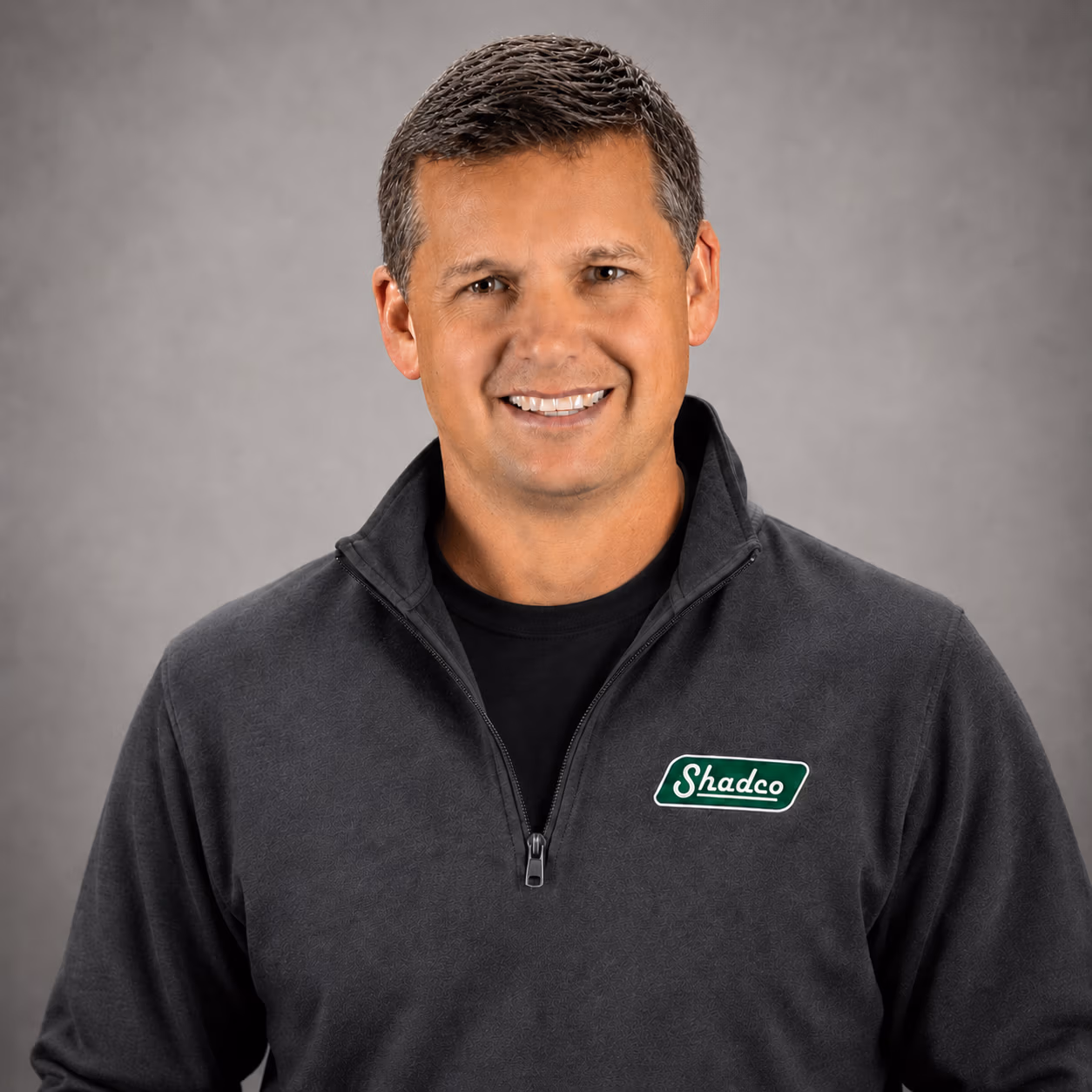 Smiling portrait of a man in a dark gray zip-up with a Shadco logo on the chest, against a gray studio backdrop.