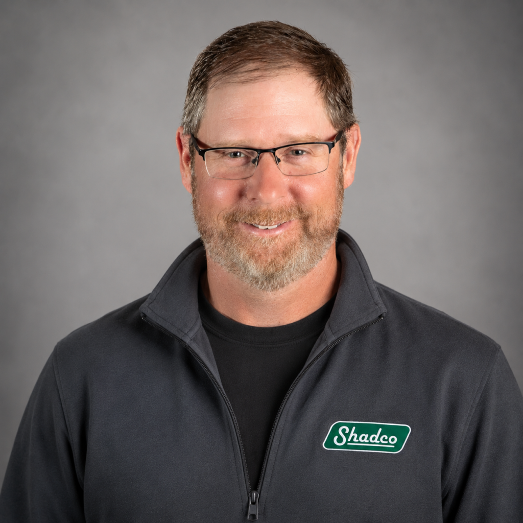 Smiling man with short brown hair and glasses, wearing a dark Shadco zip-up jacket against a gray studio background.