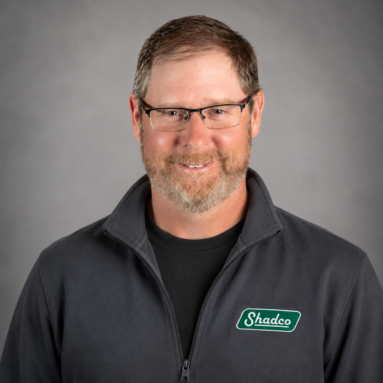 Smiling man with short brown hair and glasses, wearing a dark Shadco zip-up jacket against a gray studio background.