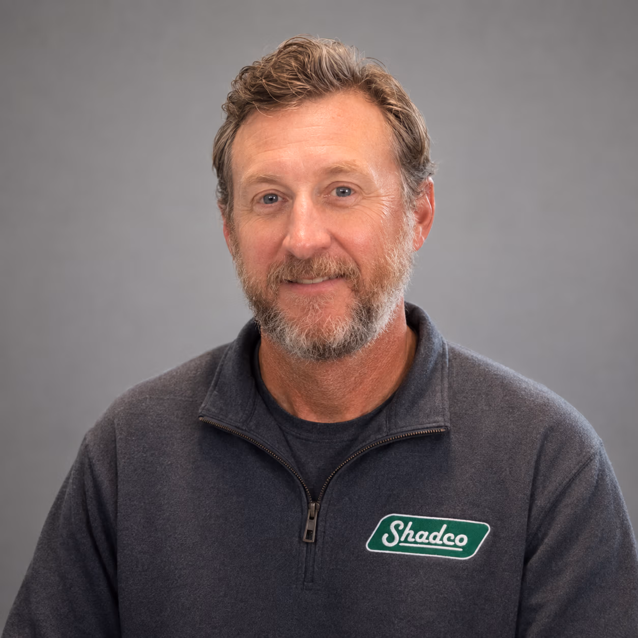 Portrait of a middle-aged man with a gray beard and short light brown hair, wearing a dark zip-up sweater with a ShadeCo logo on the chest.