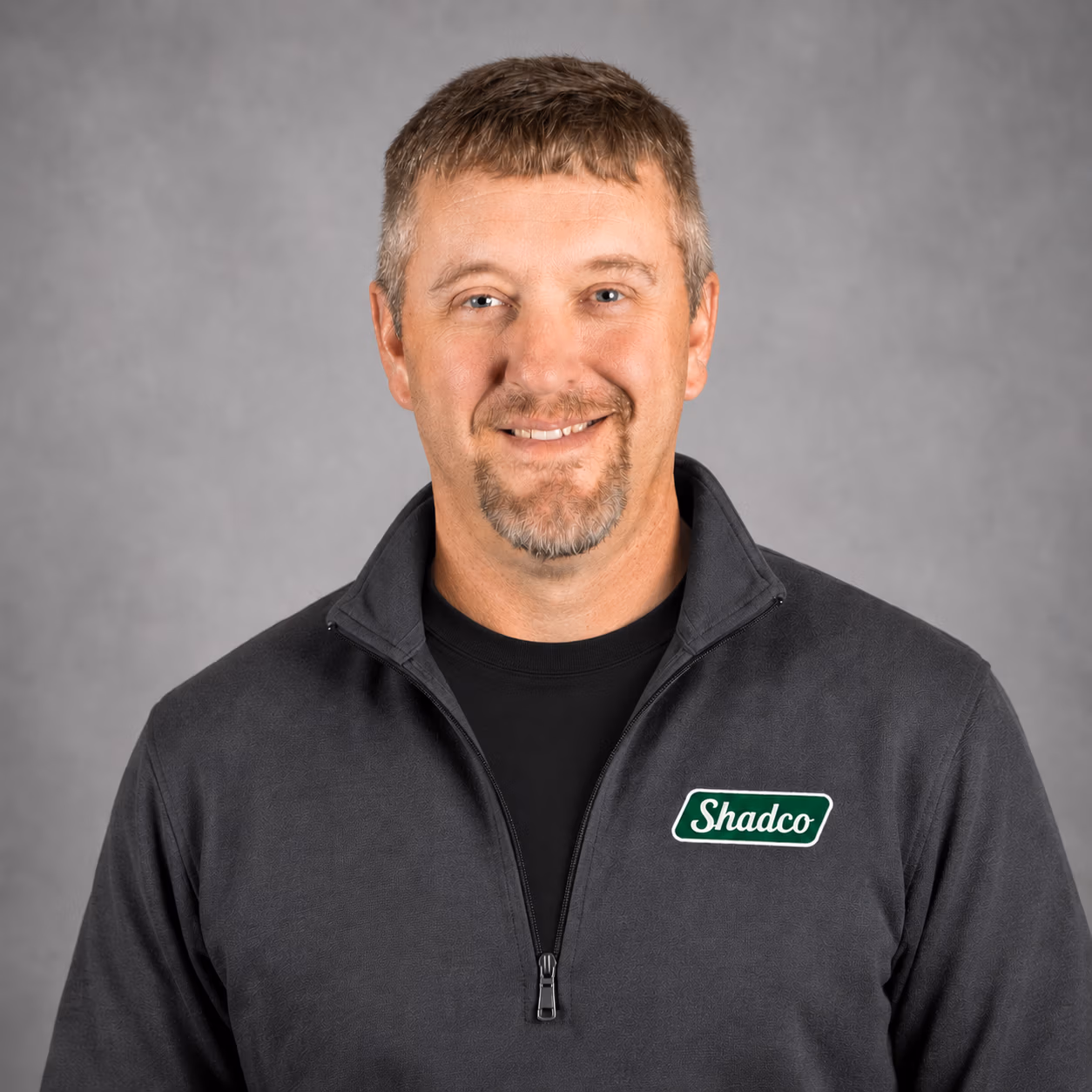 Smiling man in a dark zip-up jacket with a Shadco logo on the left chest, against a gray studio backdrop.