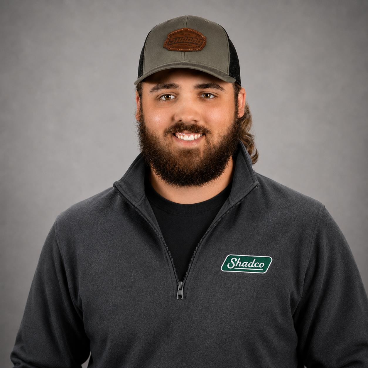Portrait of a smiling man with a beard wearing a gray quarter-zip fleece and a Shadco logo on the chest, plus a gray cap, against a neutral gray studio background.