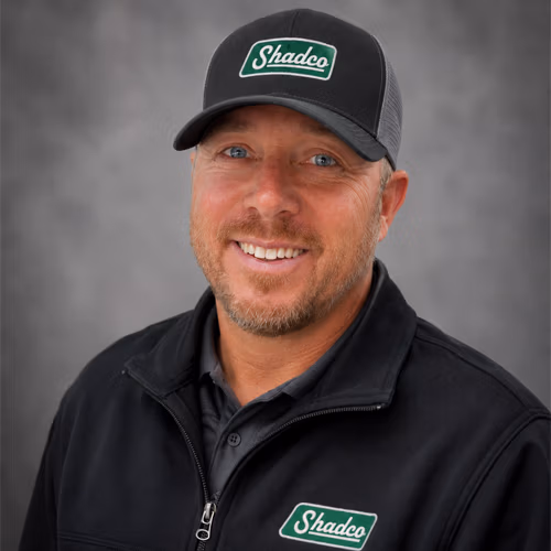 Portrait of a smiling man wearing a dark jacket and a gray Shadco cap with the logo visible on the front.