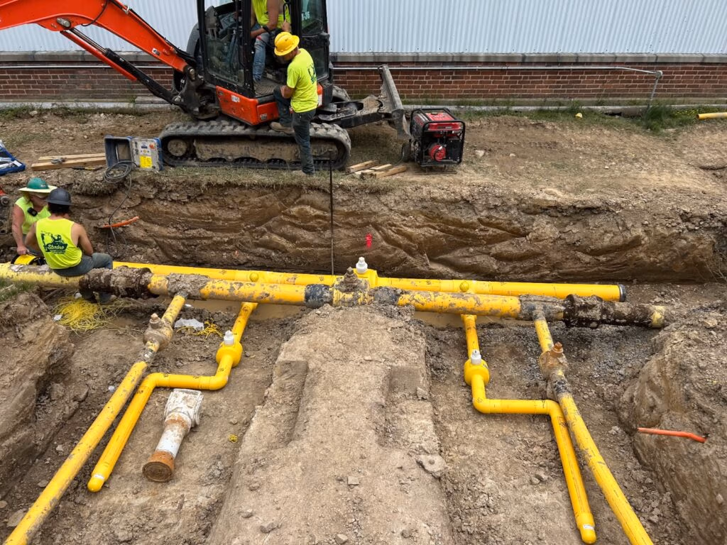 Workers in neon shirts and hard hats supervise a trench with yellow gas pipes and a small excavator above them.